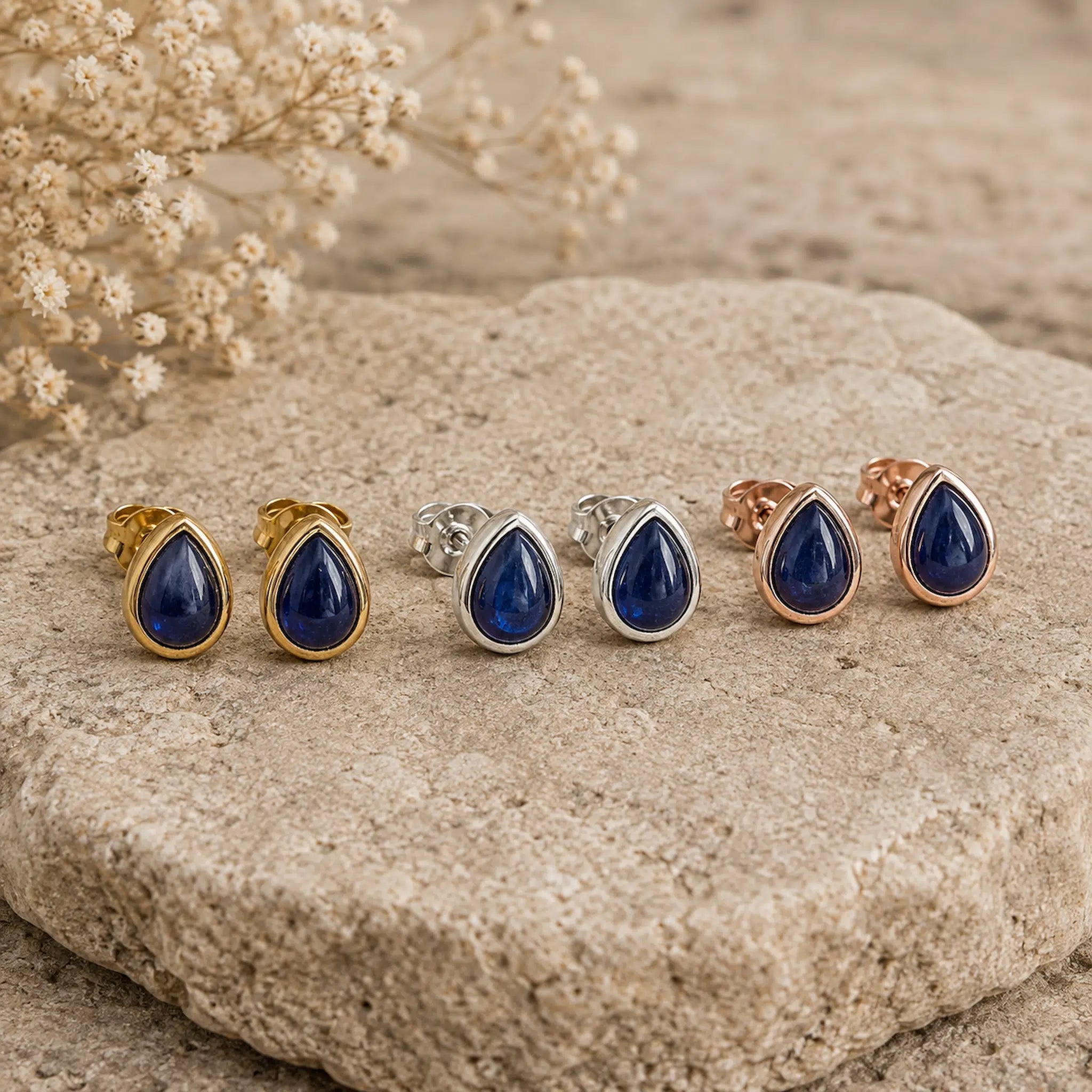 three pairs of September birthstone natural sapphire earrings - Six teardrop-shaped stud earrings with deep blue stones in gold, silver, and rose gold settings, arranged on a textured beige stone with dried flowers in the background.