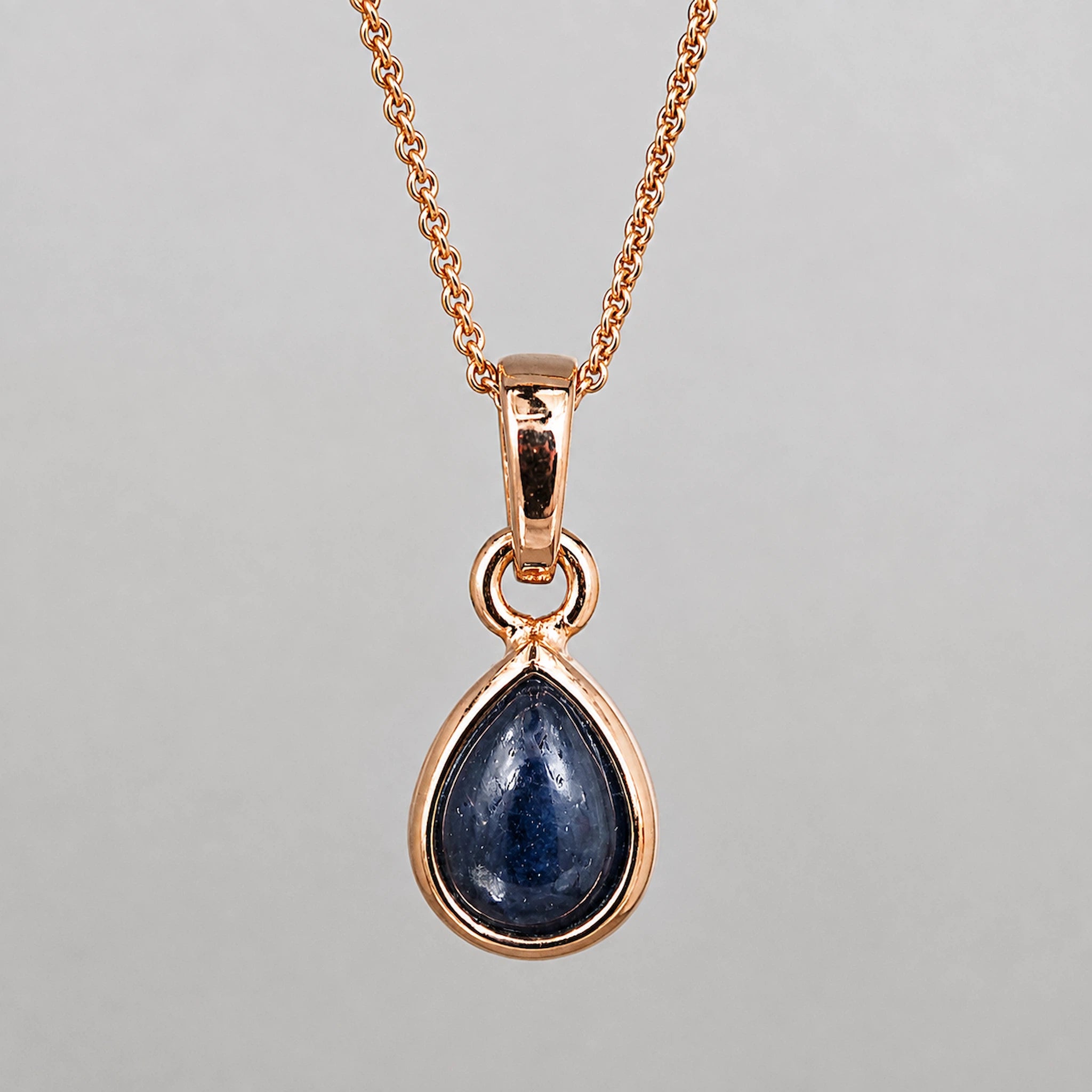 A rose gold necklace with a teardrop-shaped dark blue sapphire pendant, shown against a neutral gray background.