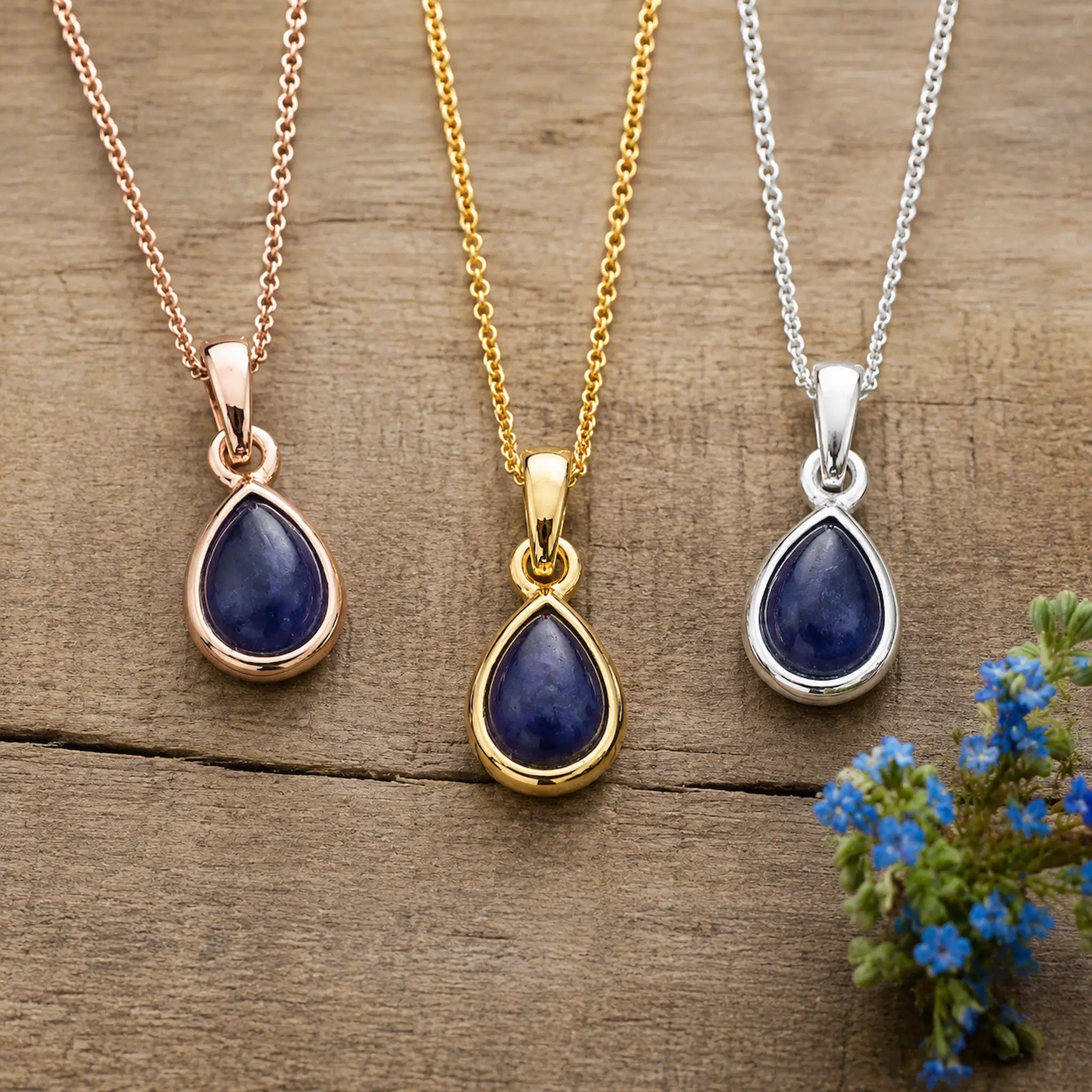 September Birthstone Natural Sapphire Necklaces - Three teardrop pendant necklaces with deep blue stones in rose gold, yellow gold, and silver settings, displayed on a rustic wooden surface with small blue flowers in the corner.