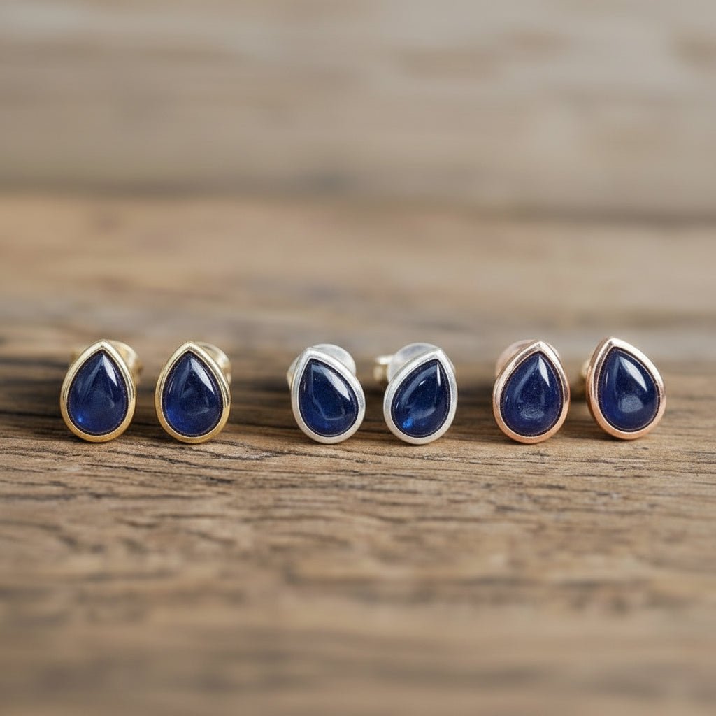 three Pairs of blue sapphire teardrop stud earrings in gold, silver and rose gold on a wooden surface