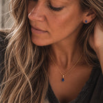 A close-up lifestyle shot of a woman wearing a september birthstone jewellery set feauring a delicate gold chain necklace with a teardrop-shaped dark blue natural sapphire gemstone pendant and matching small teardrop stud sapphire earring.