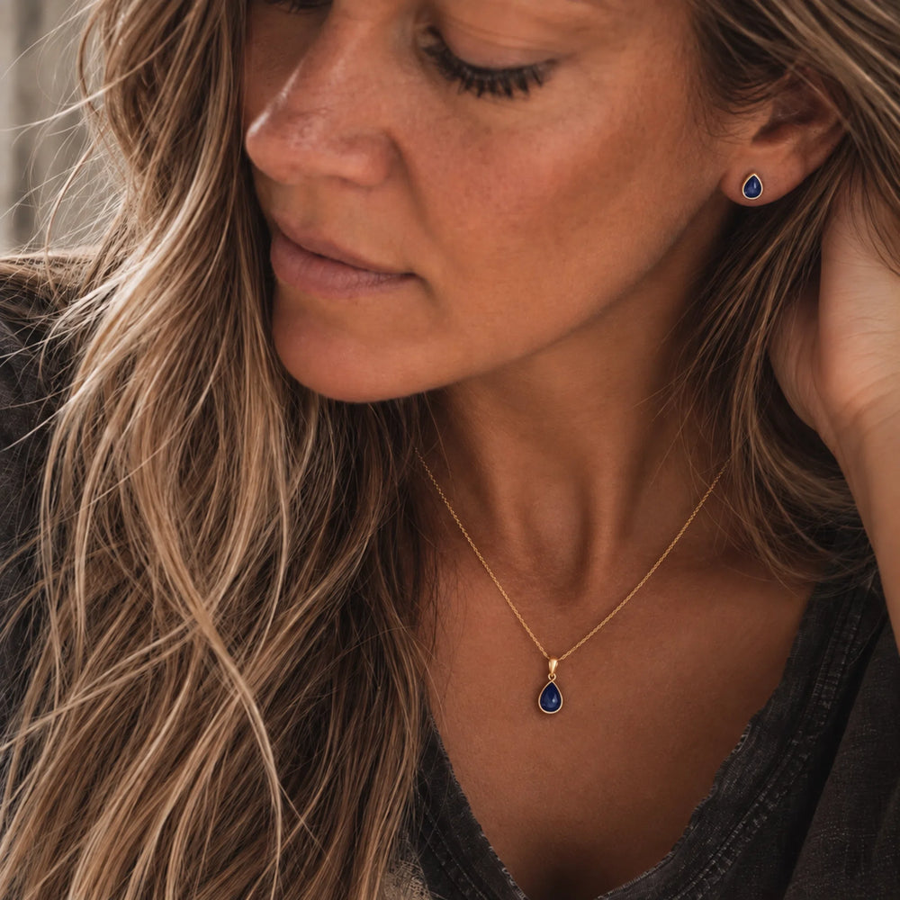 A close-up lifestyle shot of a woman wearing a september birthstone jewellery set feauring a delicate gold chain necklace with a teardrop-shaped dark blue natural sapphire gemstone pendant and matching small teardrop stud sapphire earring.