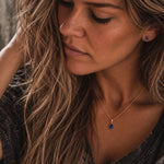 A close-up lifestyle shot of a woman wearing a  september birthstone jewellery set featuring a delicate rose gold necklace with a teardrop-shaped dark blue natural sapphire gemstone pendant, matching stud sapphire earrings, and a textured black top.