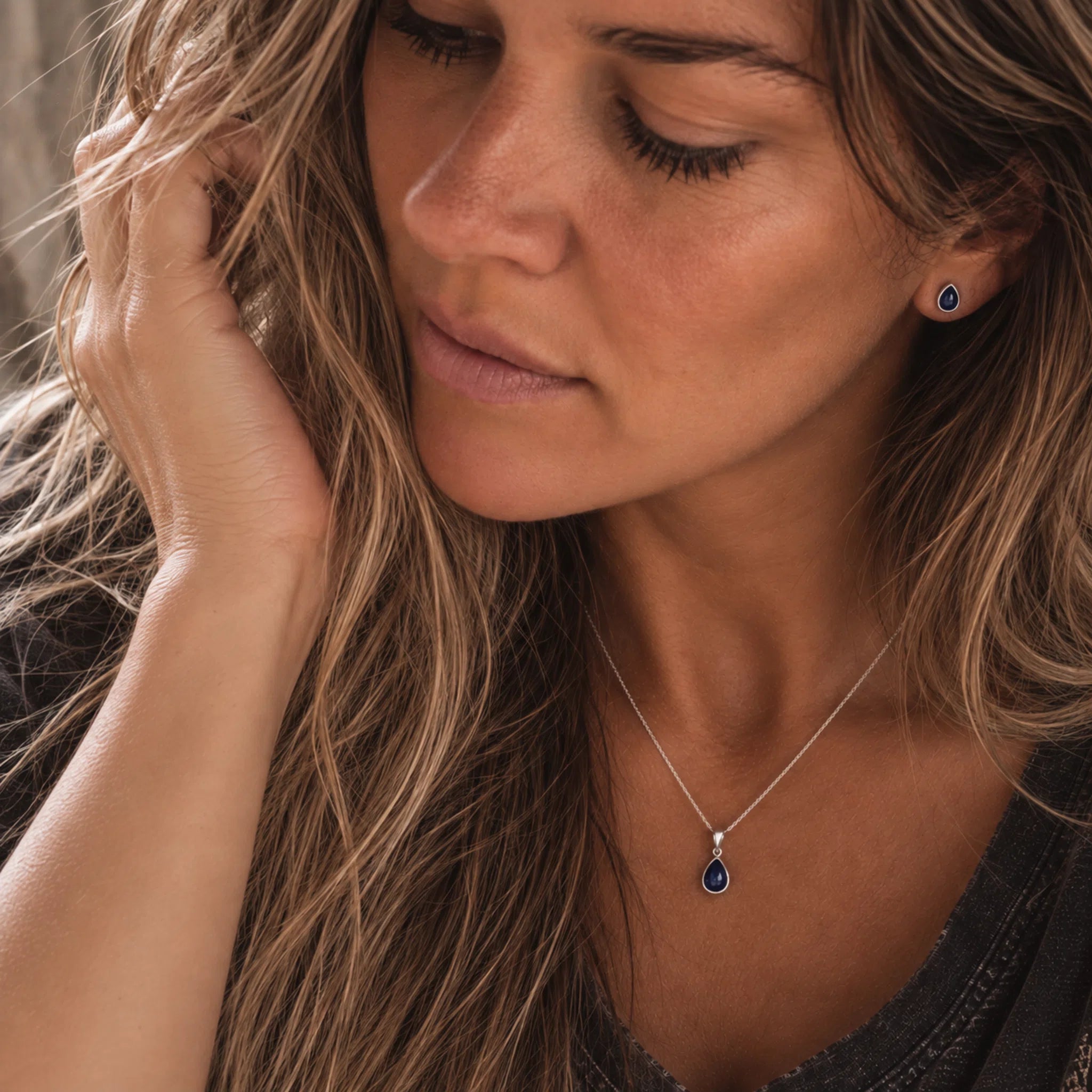 A close-up lifestyle shot of a woman wearing a sterling silver september birthstone jewellery set featuring a delicate necklace with a teardrop-shaped dark blue natural sapphire gemstone pendant and a matching stud sapphire earrings.