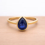 Gold ring with a blue teardrop-shaped sapphire gemstone on a wooden surface