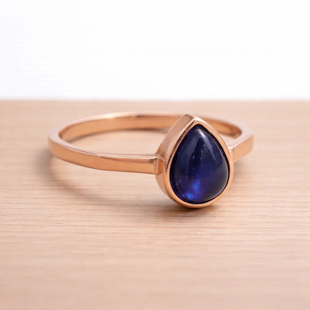 Rose gold ring with a blue teardrop sapphire stone on a beige surface