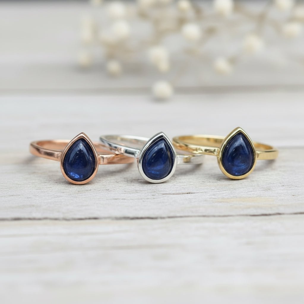 Three rings in Rose Gold, Silver and Gold with blue teardrop natural Sapphire stones on a light wooden surface.