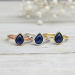Three rings in Rose Gold, Silver and Gold with blue teardrop natural Sapphire stones on a light wooden surface.