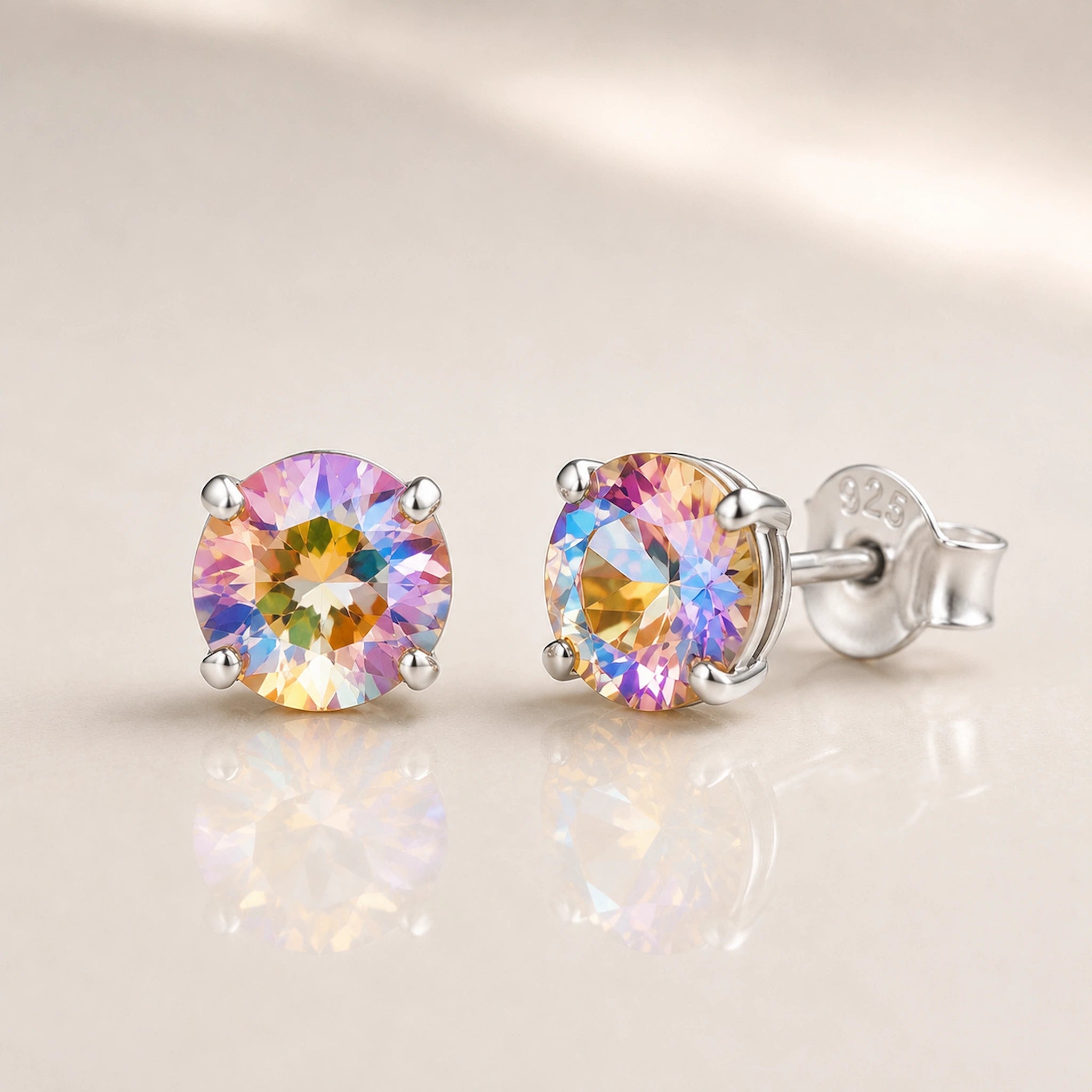A pair of round-cut silver stud earrings with iridescent, rainbow-flecked gemstones, shown on a soft beige reflective surface.