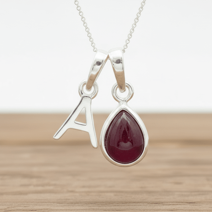 Silver necklace with a teardrop-shaped red ruby gemstone pendant and a letter 'A' pendant on a wooden surface.