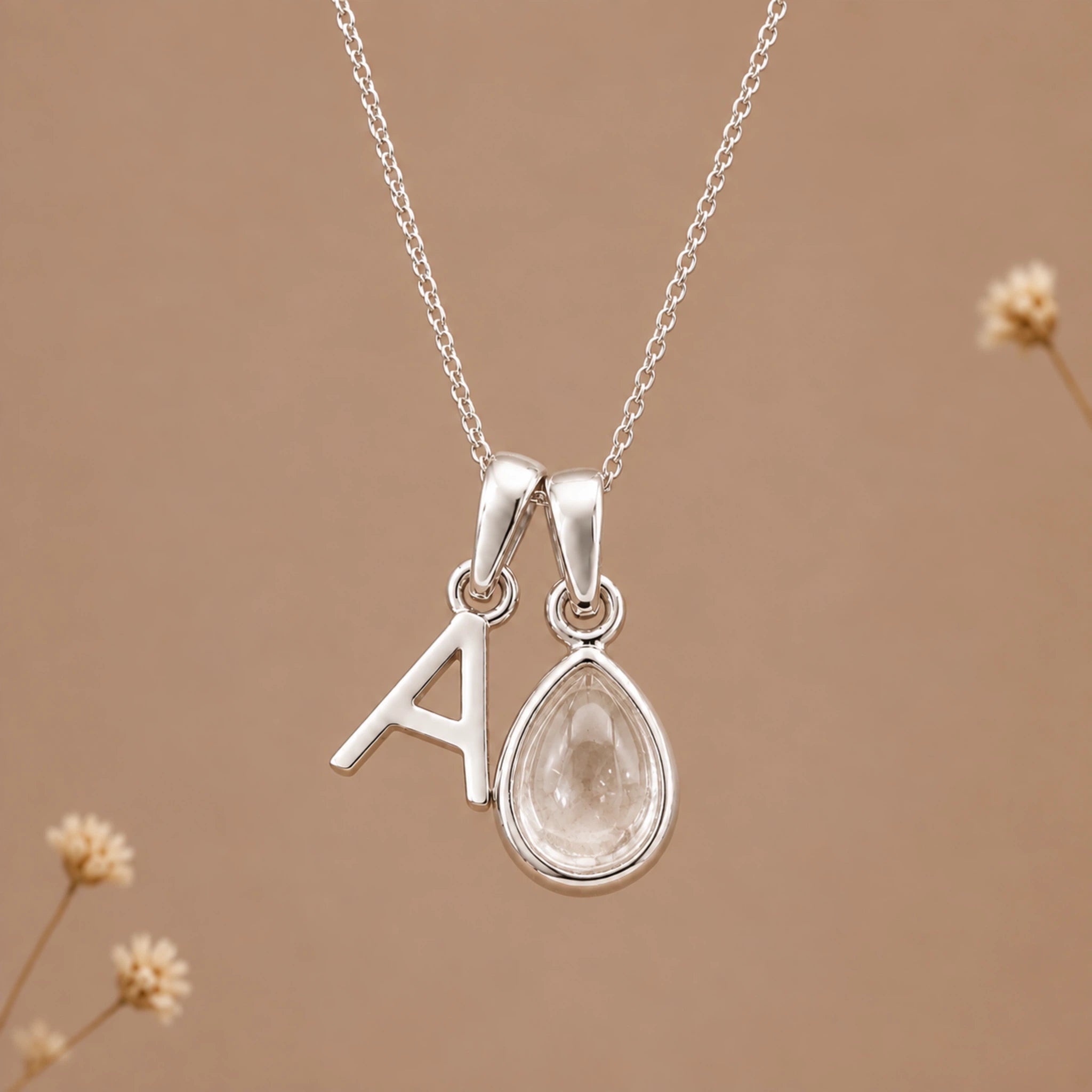 A silver necklace with a pendant featuring a letter 'A' and a teardrop-shaped clear quartz gemstone, displayed against a warm beige background with subtle dried flowers.