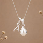 A silver necklace with two charms: a letter 'A' pendant and a teardrop-shaped pearl pendant, displayed against a warm brown background with subtle dried flower accents.