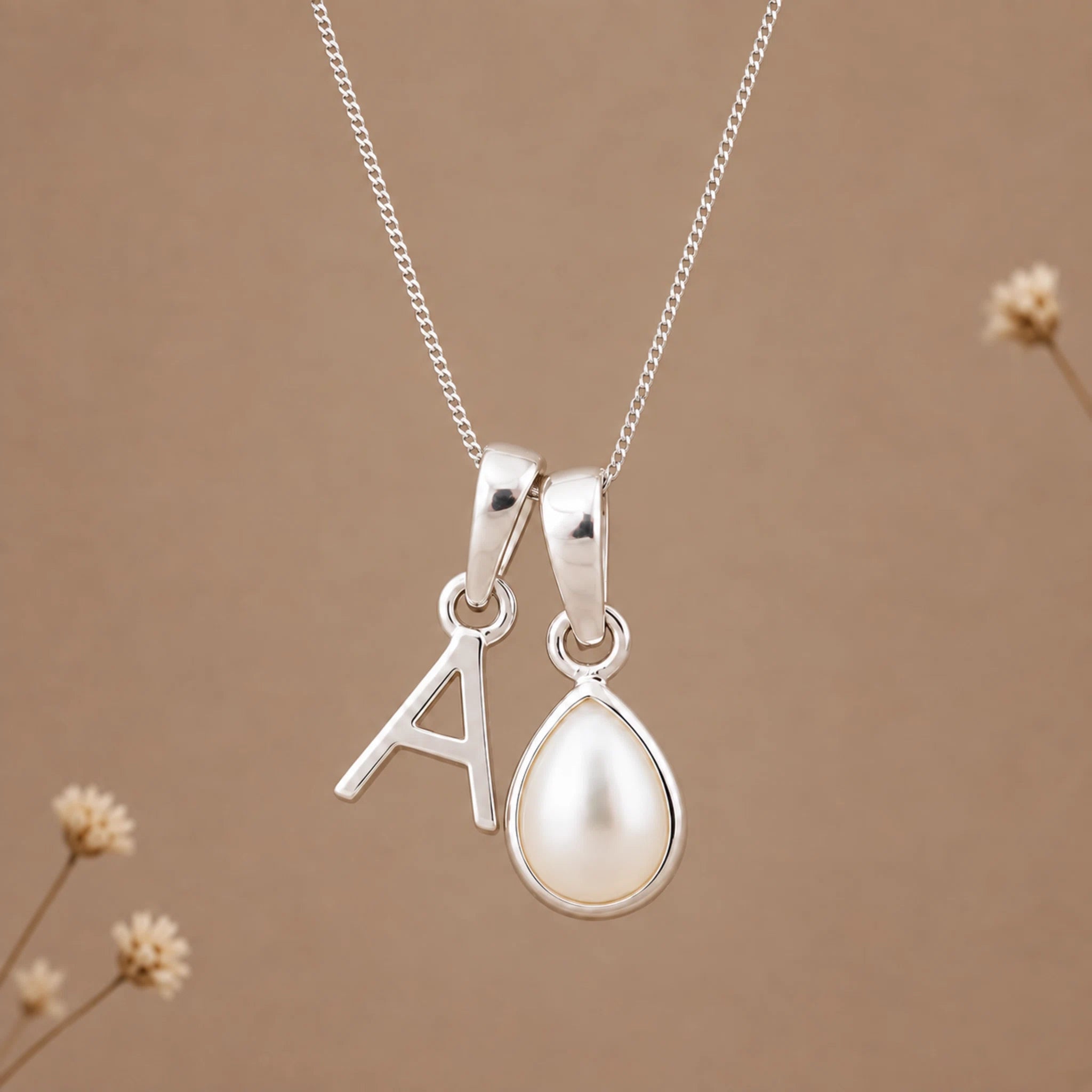 A silver necklace with two charms: a letter 'A' pendant and a teardrop-shaped pearl pendant, displayed against a warm brown background with subtle dried flower accents.