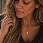 A close-up lifestyle shot of a woman wearing a delicate silver necklace with a pear-shaped pearl pendant. The necklace features a silver bail with two small letter charms, one of which appears to spell 'A'.
