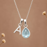 sterling silver initial pendant necklace featuring the letter 'A' and a march birthstone teardrop-shaped natural aquamarine gemstone charm, displayed against a warm beige background with subtle dried flowers.