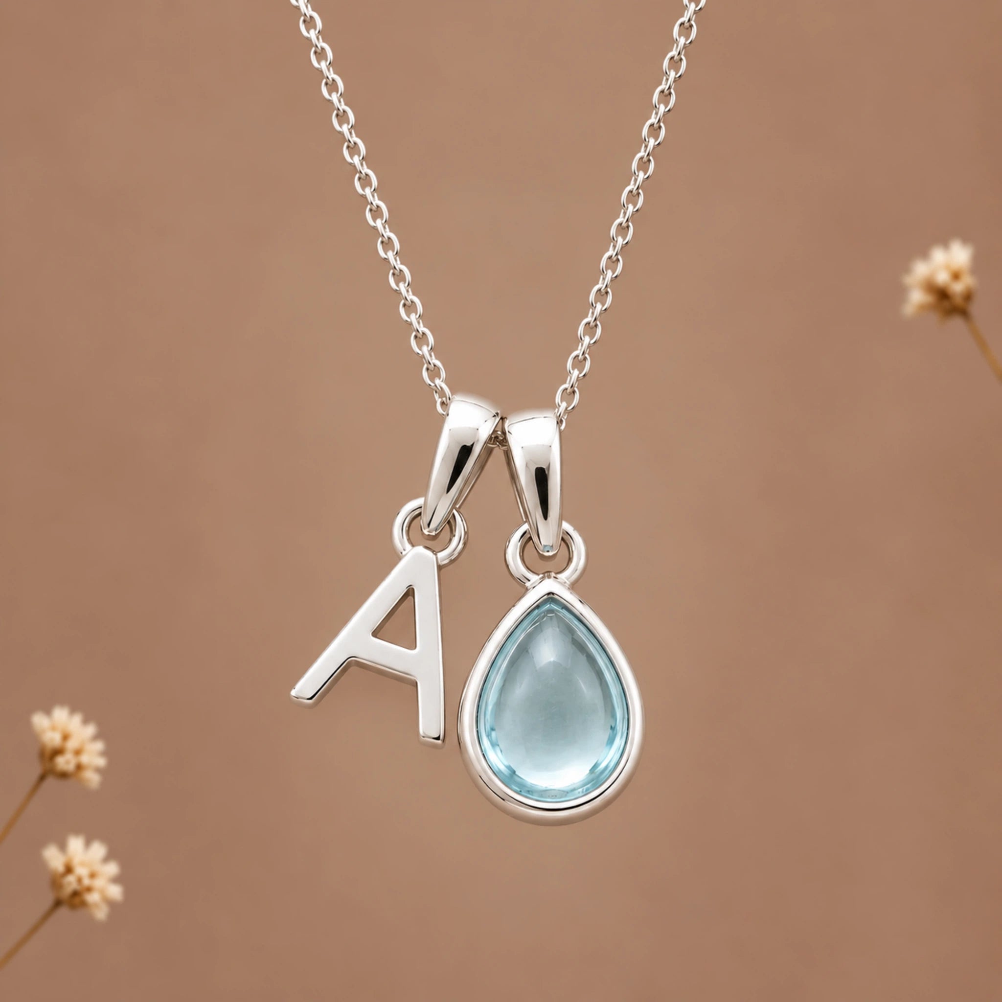 sterling silver initial pendant necklace featuring the letter 'A' and a march birthstone teardrop-shaped natural aquamarine gemstone charm, displayed against a warm beige background with subtle dried flowers.