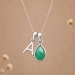 A silver initial necklace featuring a letter 'A' pendant and a teardrop-shaped green gemstone pendant, displayed on a soft beige background with subtle dried flower accents.