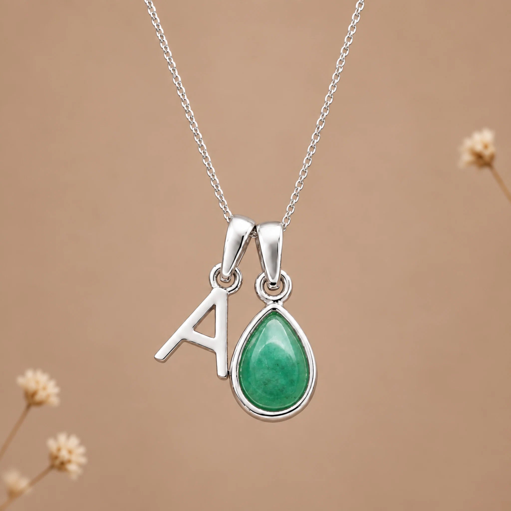 A silver initial necklace featuring a letter 'A' pendant and a teardrop-shaped green gemstone pendant, displayed on a soft beige background with subtle dried flower accents.