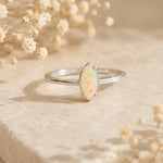 A silver band ring featuring a marquise-cut opal gemstone with iridescent white, pink, and blue flashes, displayed on a textured beige stone with dried baby's breath flowers.