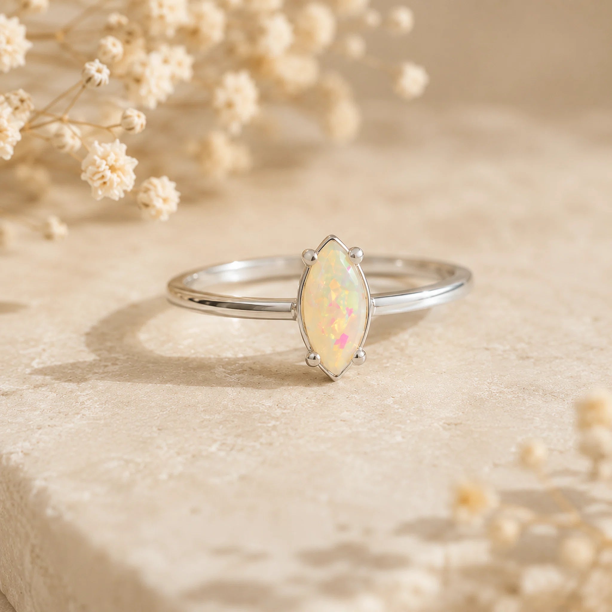 A silver band ring featuring a marquise-cut opal gemstone with iridescent white, pink, and blue flashes, displayed on a textured beige stone with dried baby's breath flowers.