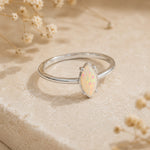 A silver ring featuring a marquise-cut white opal gemstone, displayed on a textured beige stone surface with dried flowers in the background.
