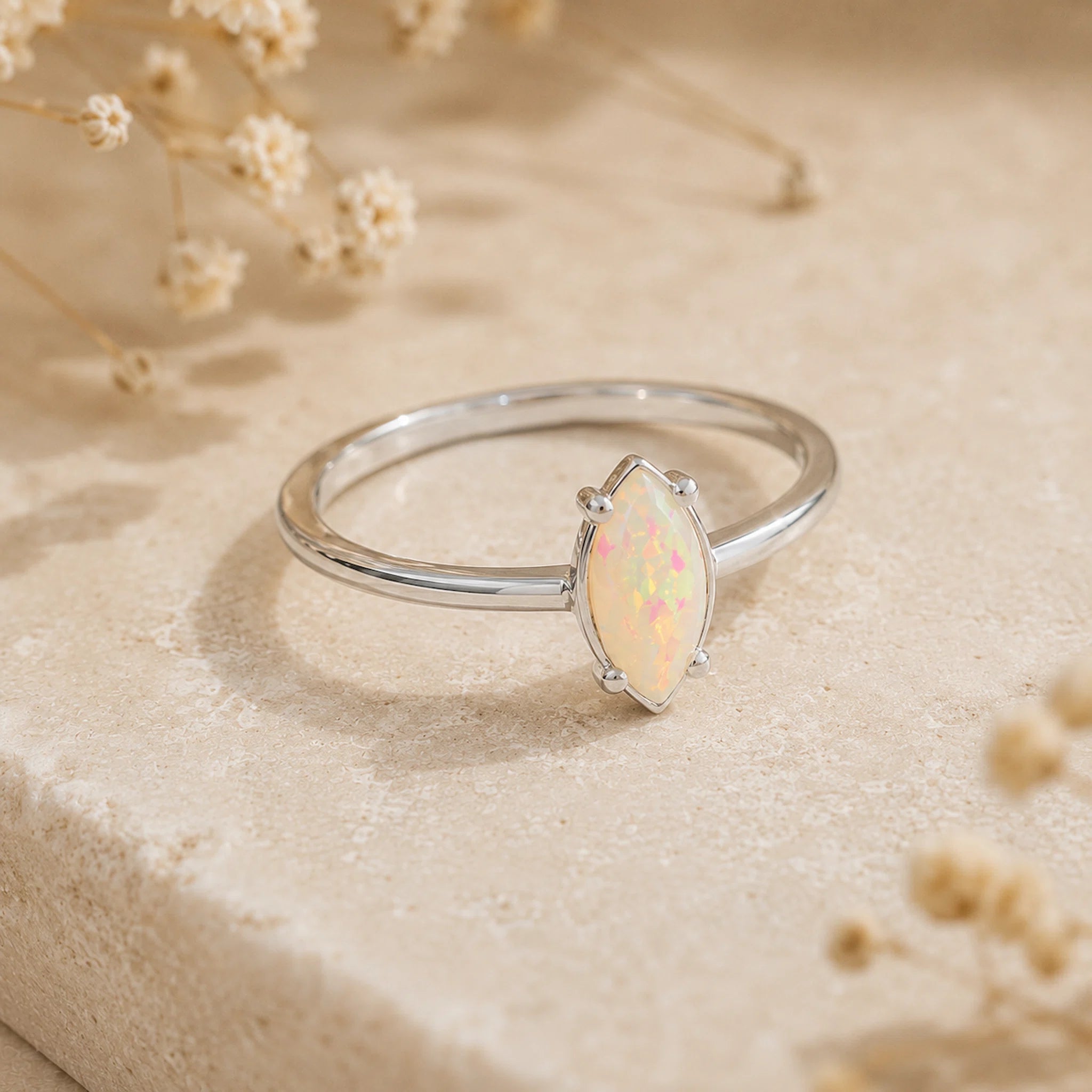 A silver ring featuring a marquise-cut white opal gemstone, displayed on a textured beige stone surface with dried flowers in the background.