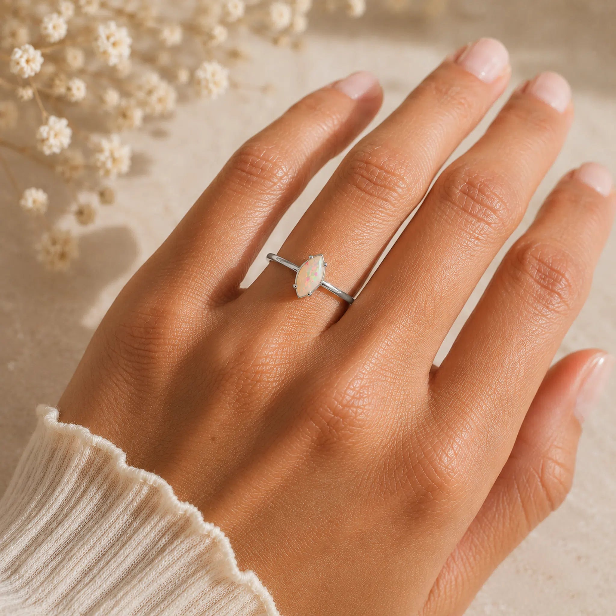 A silver ring with a marquise-cut white stone worn on a hand, styled with dried white flowers in the background.