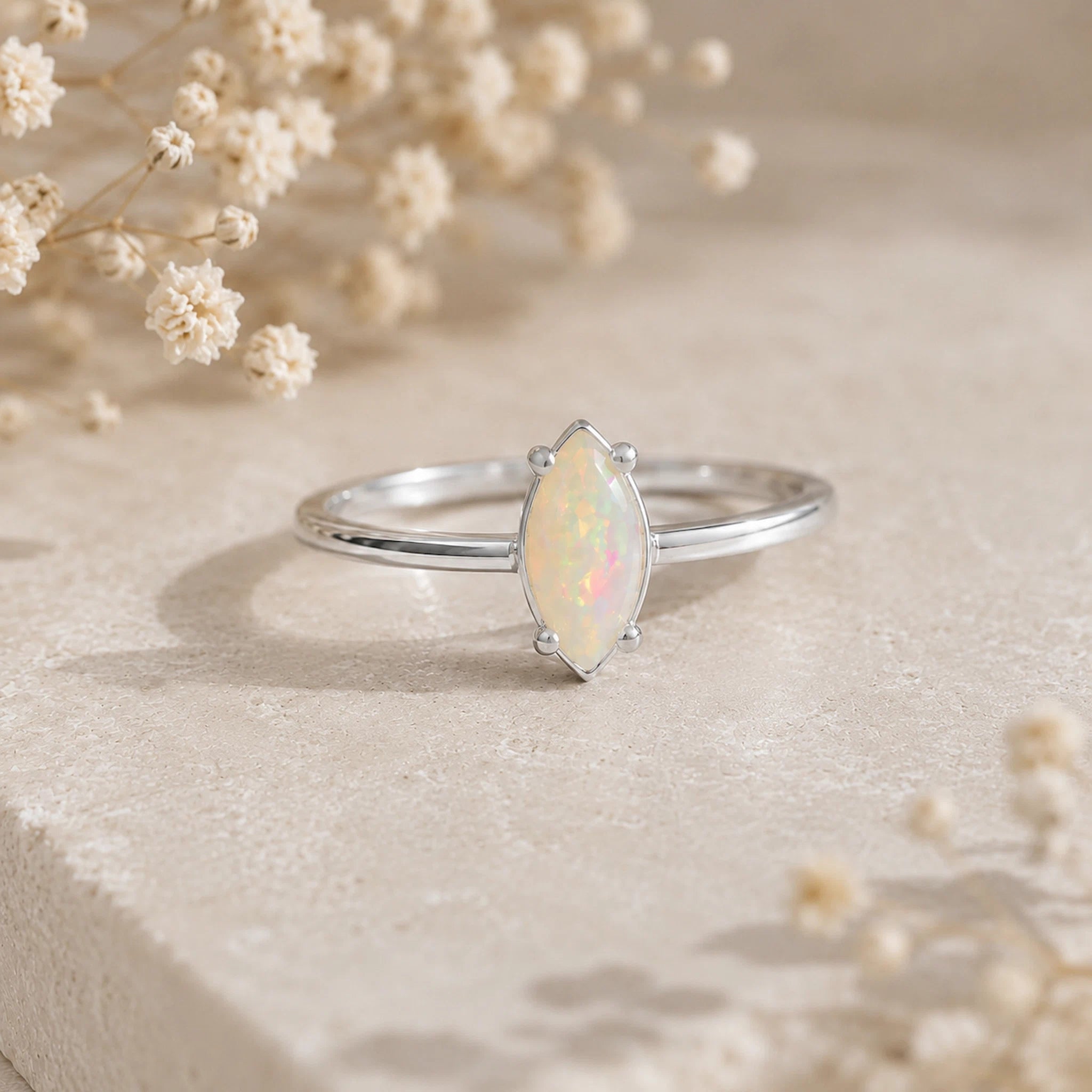 A silver ring with a marquise-cut white opal center stone, displayed on a textured beige stone surface with dried baby's breath flowers in the background.