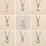 Collection of silver letter necklaces J to Q on a beige background as initial options for a personalised April Birthstone Clear Quartz Necklace.