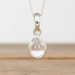 Teardrop-shaped pendant with a clear stone on a chain against a neutral background