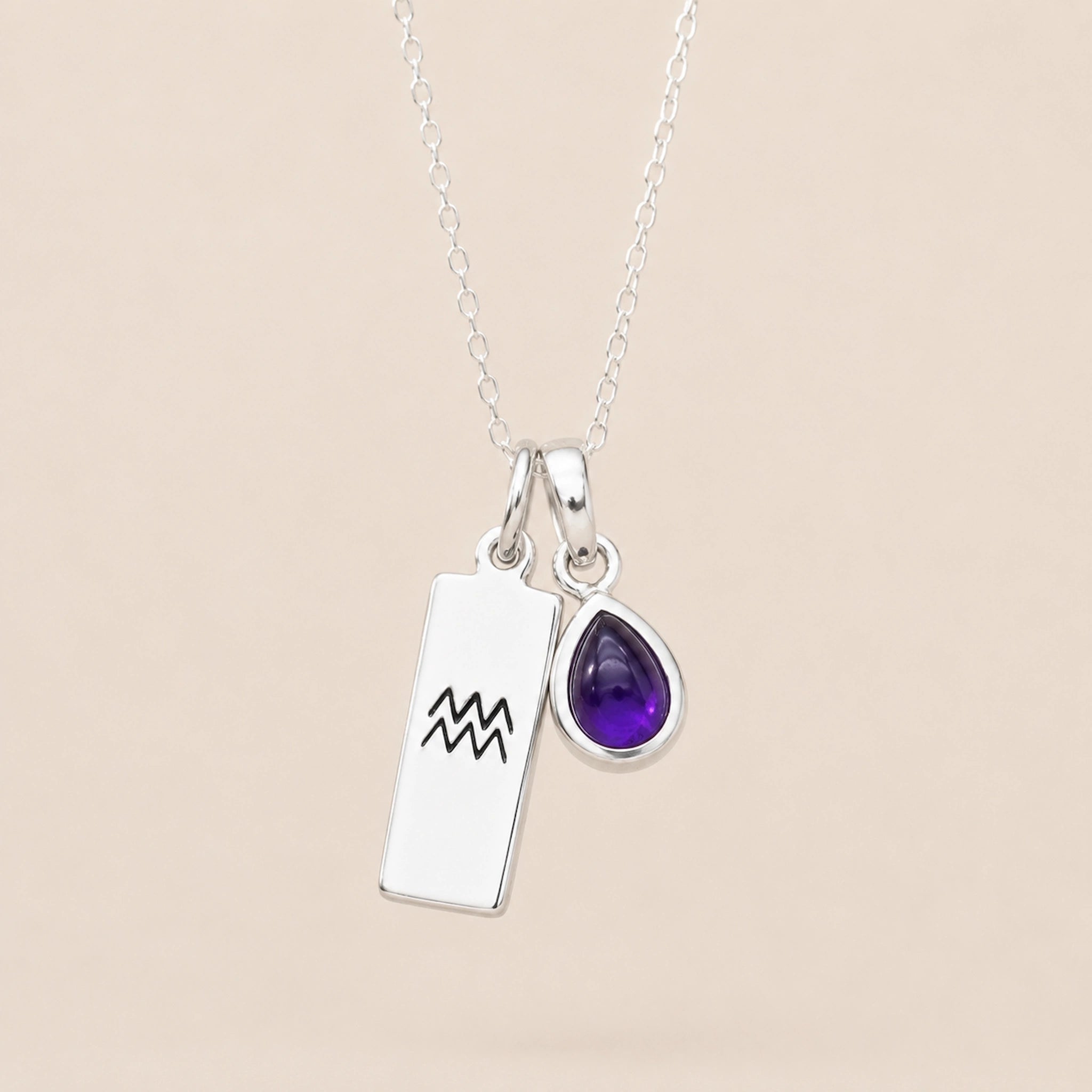 Sterling Silver necklace with a teardrop february birthstone natural amethyst pendant and engraved aquarius glyph tag hanging on a beige background.