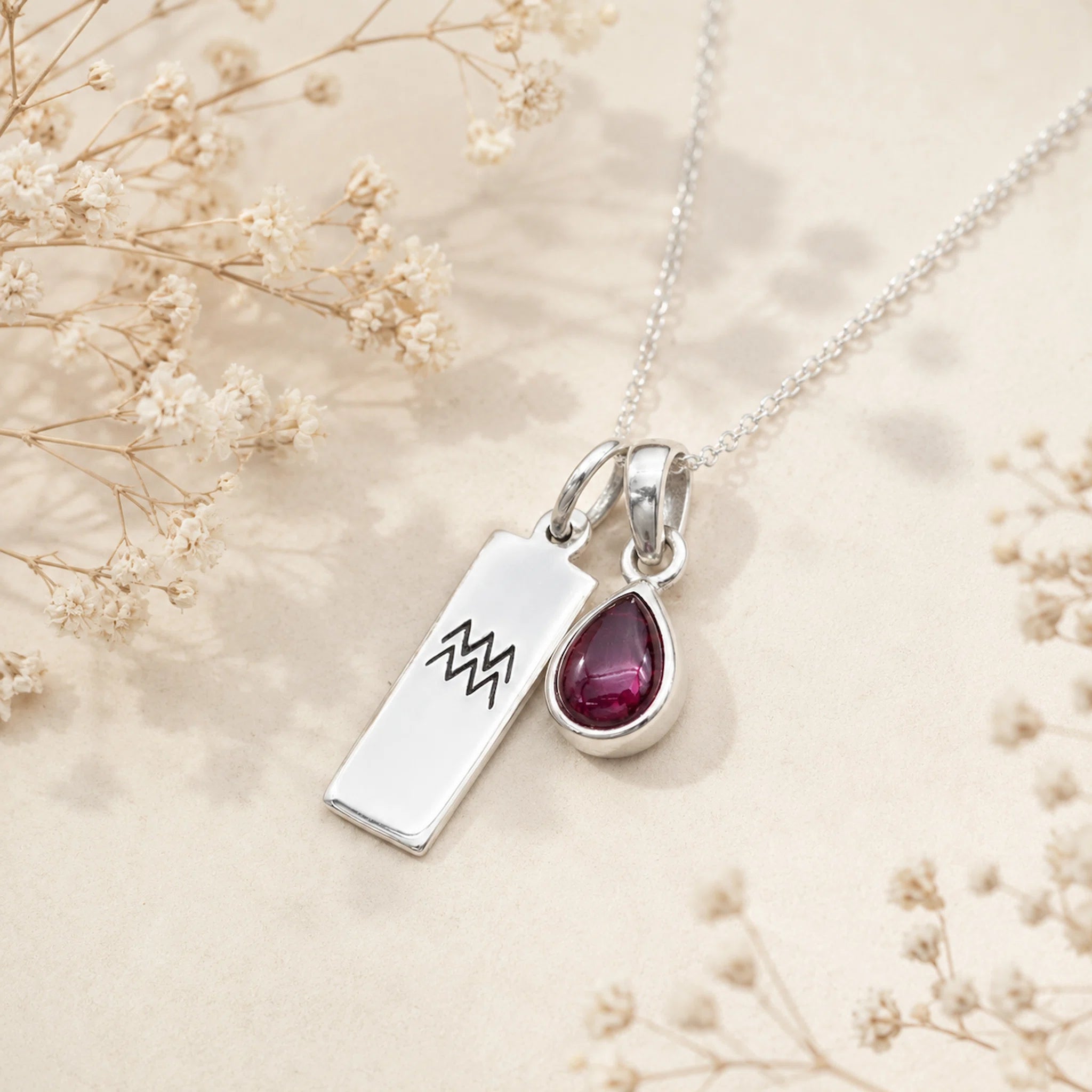 Sterling Silver necklace with a teardrop january birthstone natural garnet pendant and engraved aquarius glyph tag on a beige background with dried flowers.