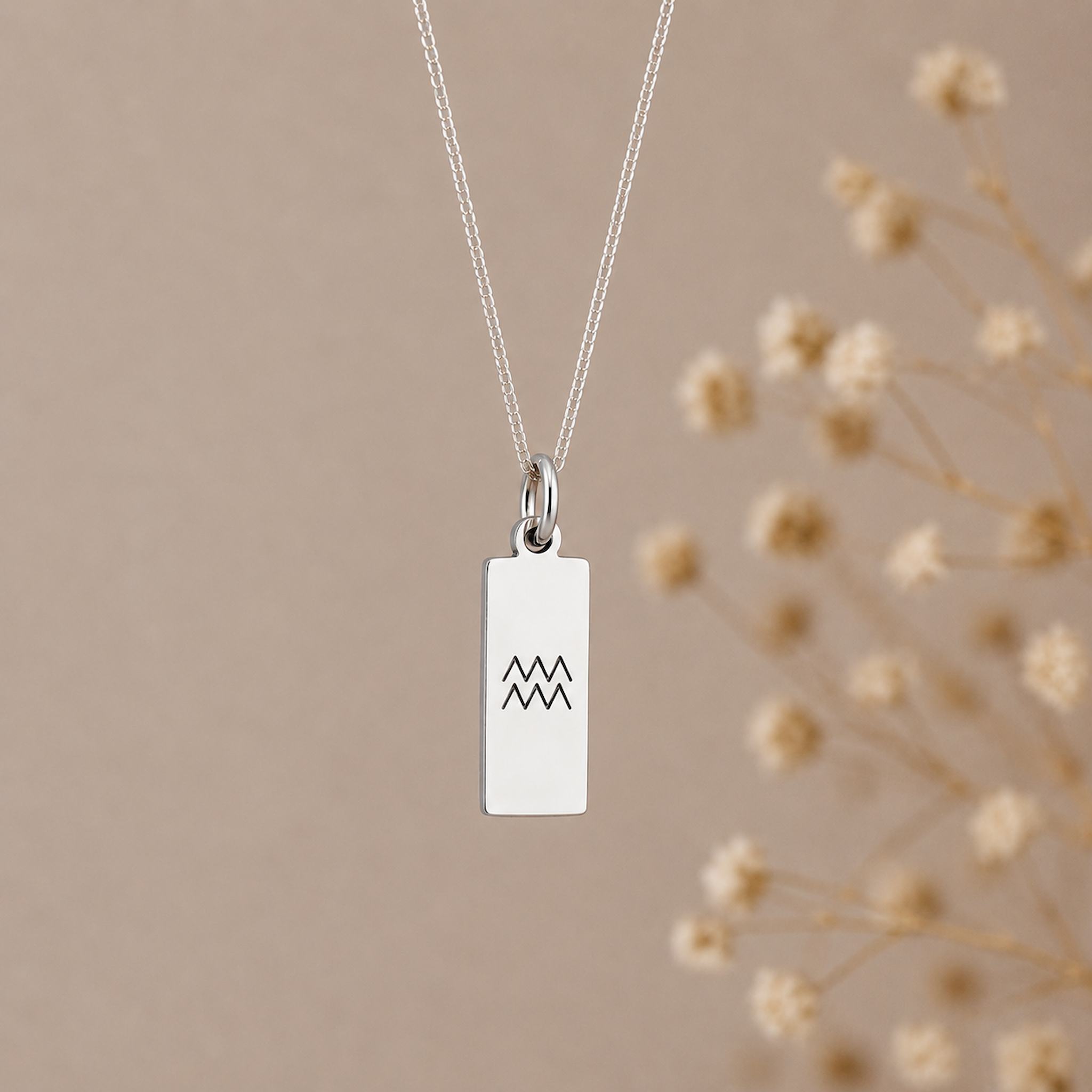 aquarius necklace - A silver rectangular pendant necklace with a black engraved aquarius star sign line glyph symbol, hanging from a delicate silver chain against a warm beige background 