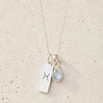 Sterling silver necklace with march birthstone aquamarine pendant and rectangle pendant with engraved aries line glyph hanging on a light background.