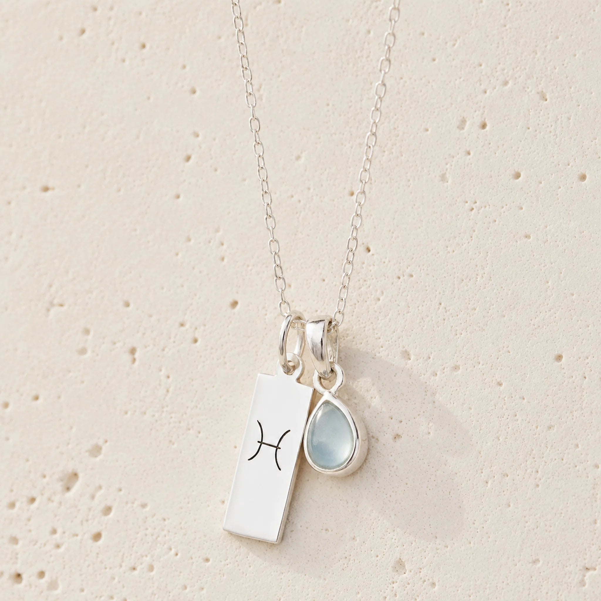 Sterling silver necklace with march birthstone aquamarine pendant and rectangle pendant with engraved aries line glyph hanging on a light background.