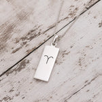 A silver rectangular pendant necklace engraved with the Aries zodiac symbol, resting on a rustic, light-colored wooden surface.