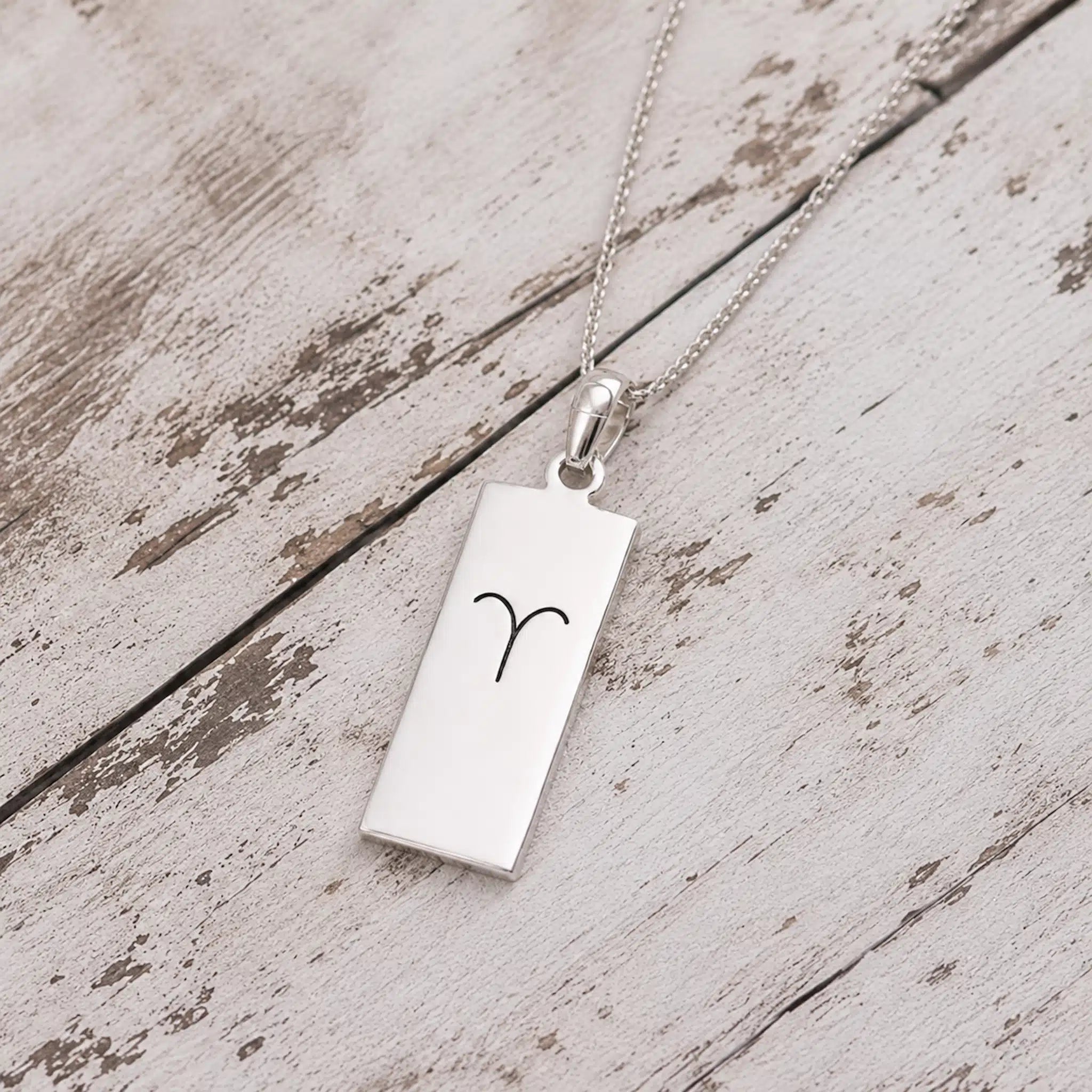 A silver rectangular pendant necklace engraved with the Aries zodiac symbol, resting on a rustic, light-colored wooden surface.
