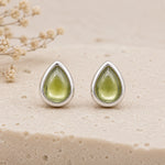 A pair of August Birthstone Natural Peridot Earrings - silver teardrop stud earrings with smooth, polished light green gemstone centers, displayed on a textured beige stone surface with dried flowers in the background.