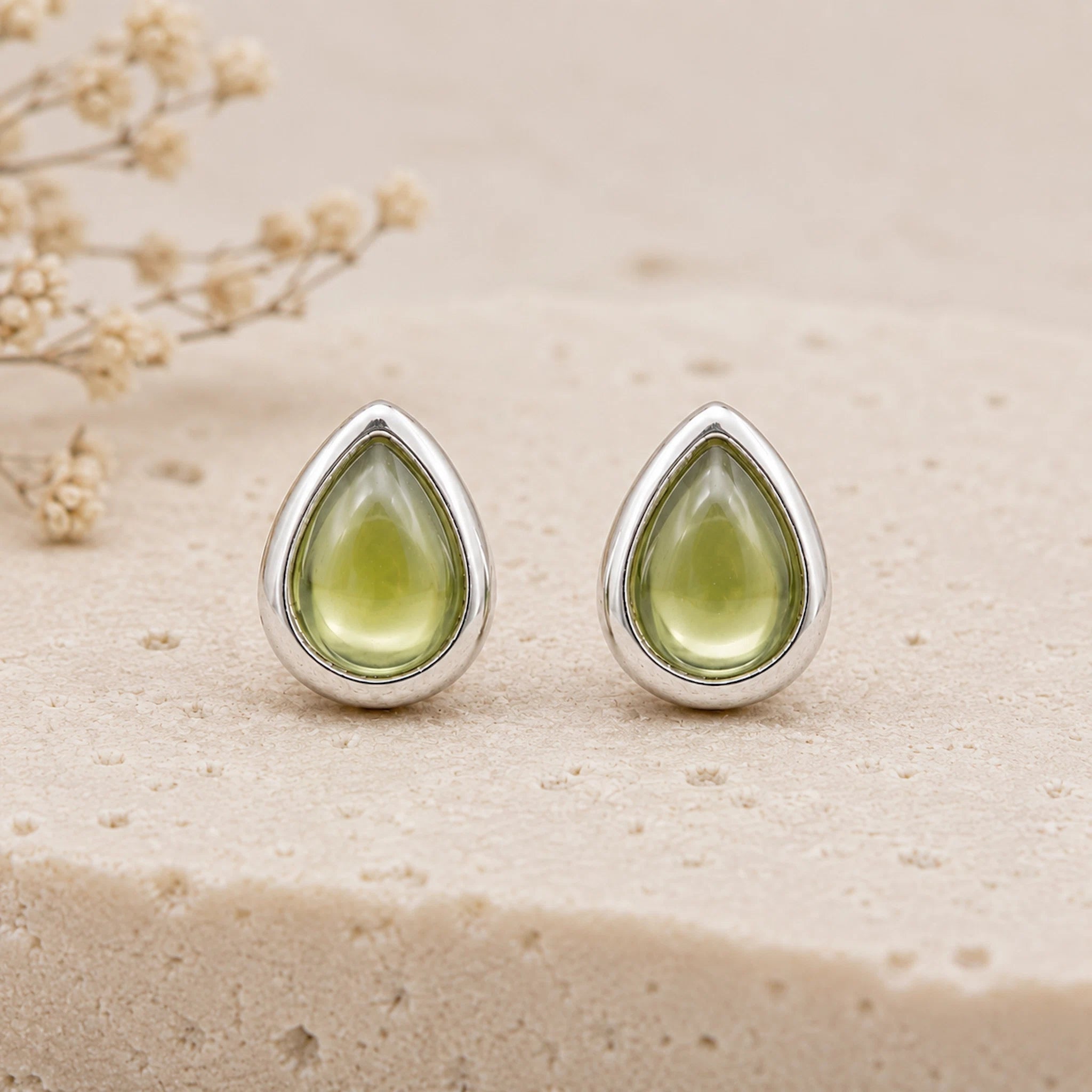 A pair of August Birthstone Natural Peridot Earrings - silver teardrop stud earrings with smooth, polished light green gemstone centers, displayed on a textured beige stone surface with dried flowers in the background.