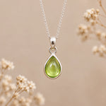A silver august birthstone necklace featuring a teardrop pendant with a polished green natural peridot gemstone, suspended from a delicate silver chain against a warm beige background with soft blurred dried flowers.