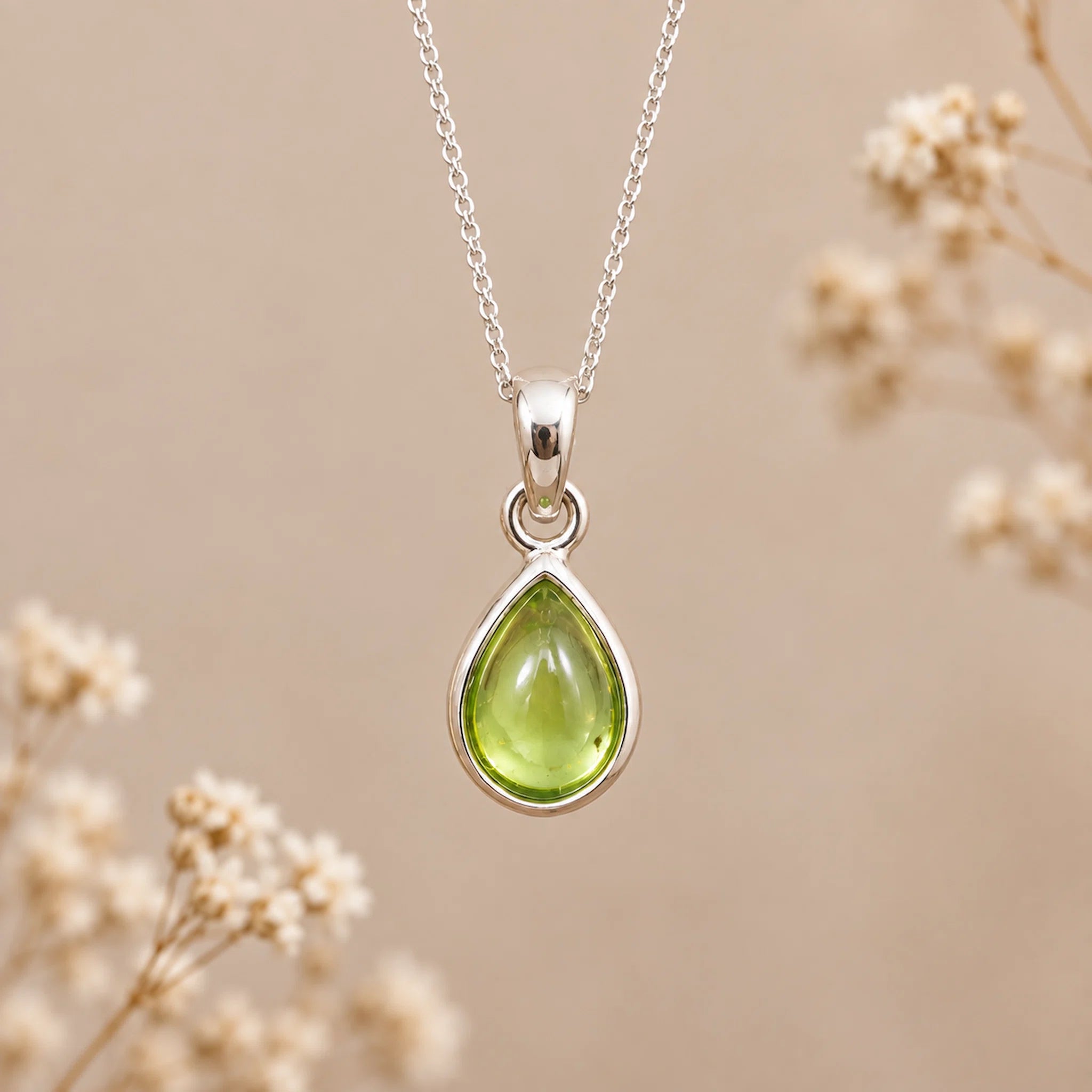 A silver august birthstone necklace featuring a teardrop pendant with a polished green natural peridot gemstone, suspended from a delicate silver chain against a warm beige background with soft blurred dried flowers.