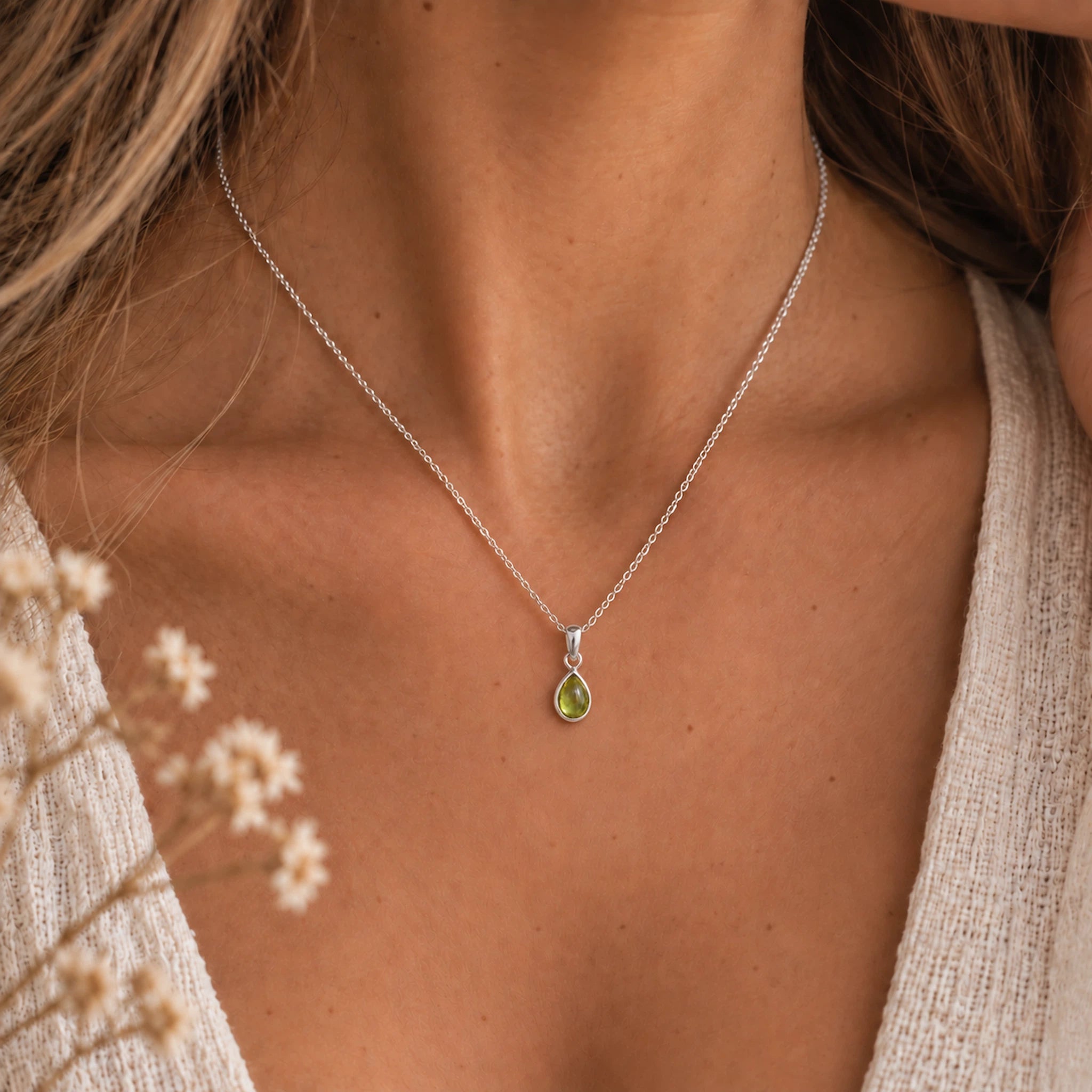 A close-up of a woman's neck and chest wearing a august birthstone necklace featuring a delicate silver chain necklace with a small teardrop-shaped green peridot gemstone pendant