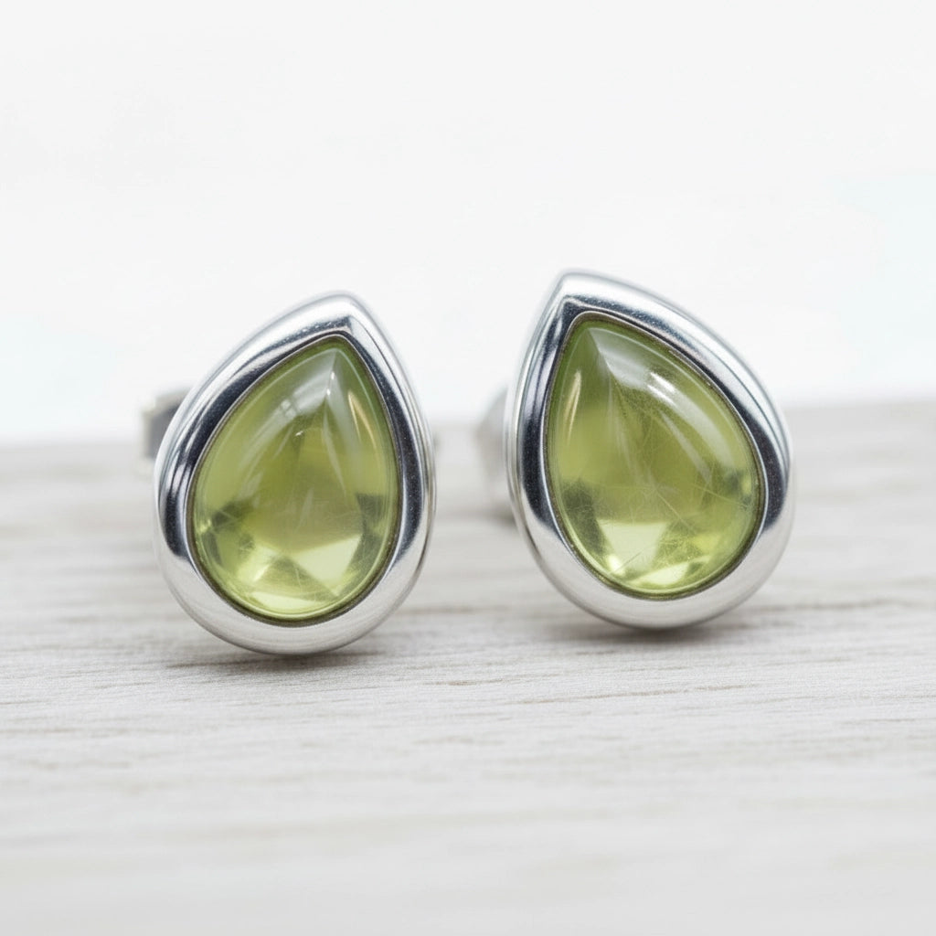 Teardrop-shaped green peridot gemstone stud earrings with silver settings on a light wooden surface.
