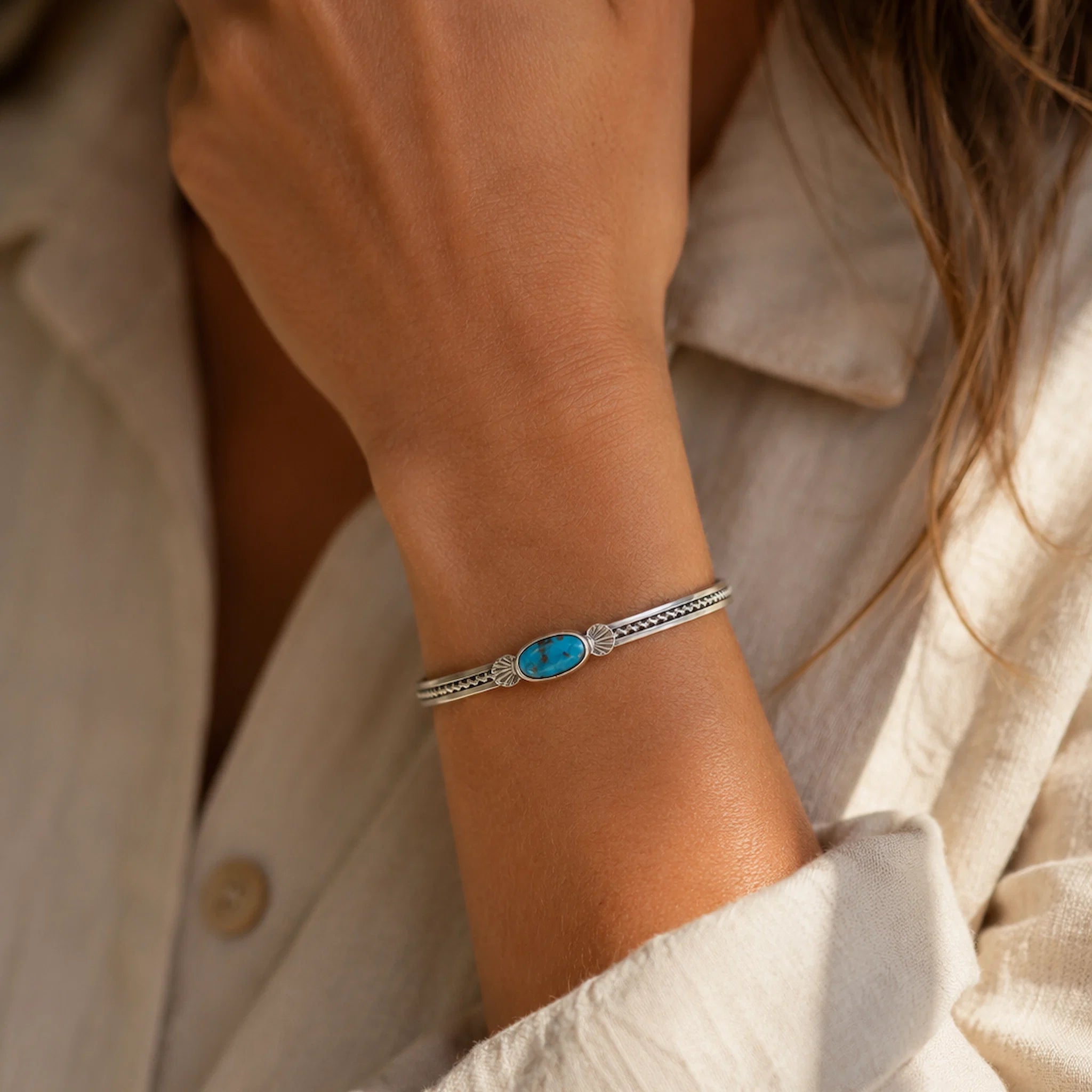 A close-up of a person's wrist wearing a silver-toned cuff bracelet with a central oval turquoise-blue stone and small clear stones on either side, paired with a light beige linen shirt.