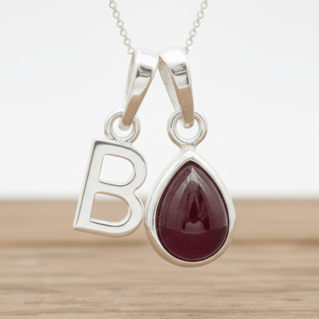 Silver necklace with a 'B' pendant and a teardrop-shaped red ruby gemstone on a wooden surface.