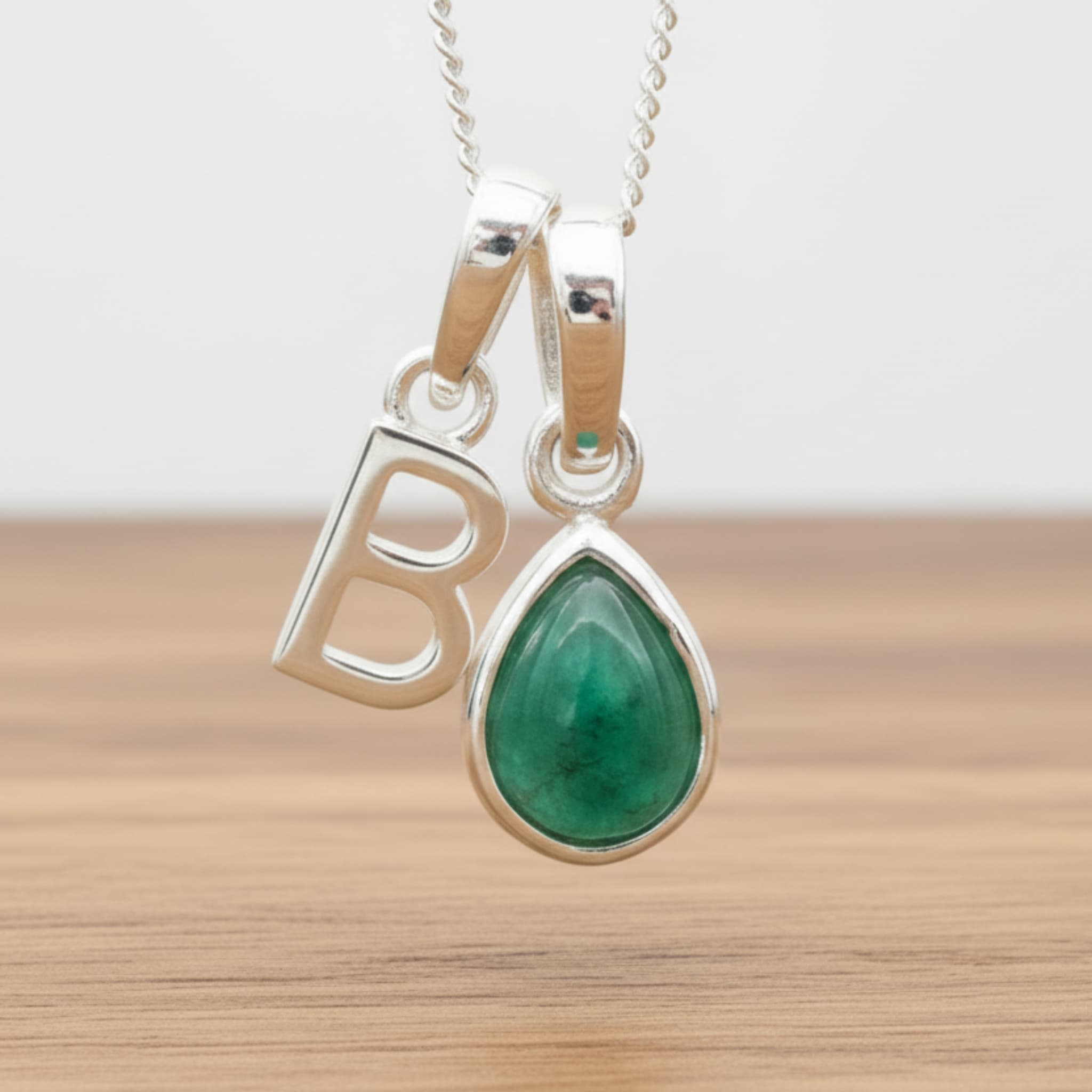 Silver Initial B necklace with a teardrop-shaped Emerald gemstone pendant hanging above a rustic wooden surface