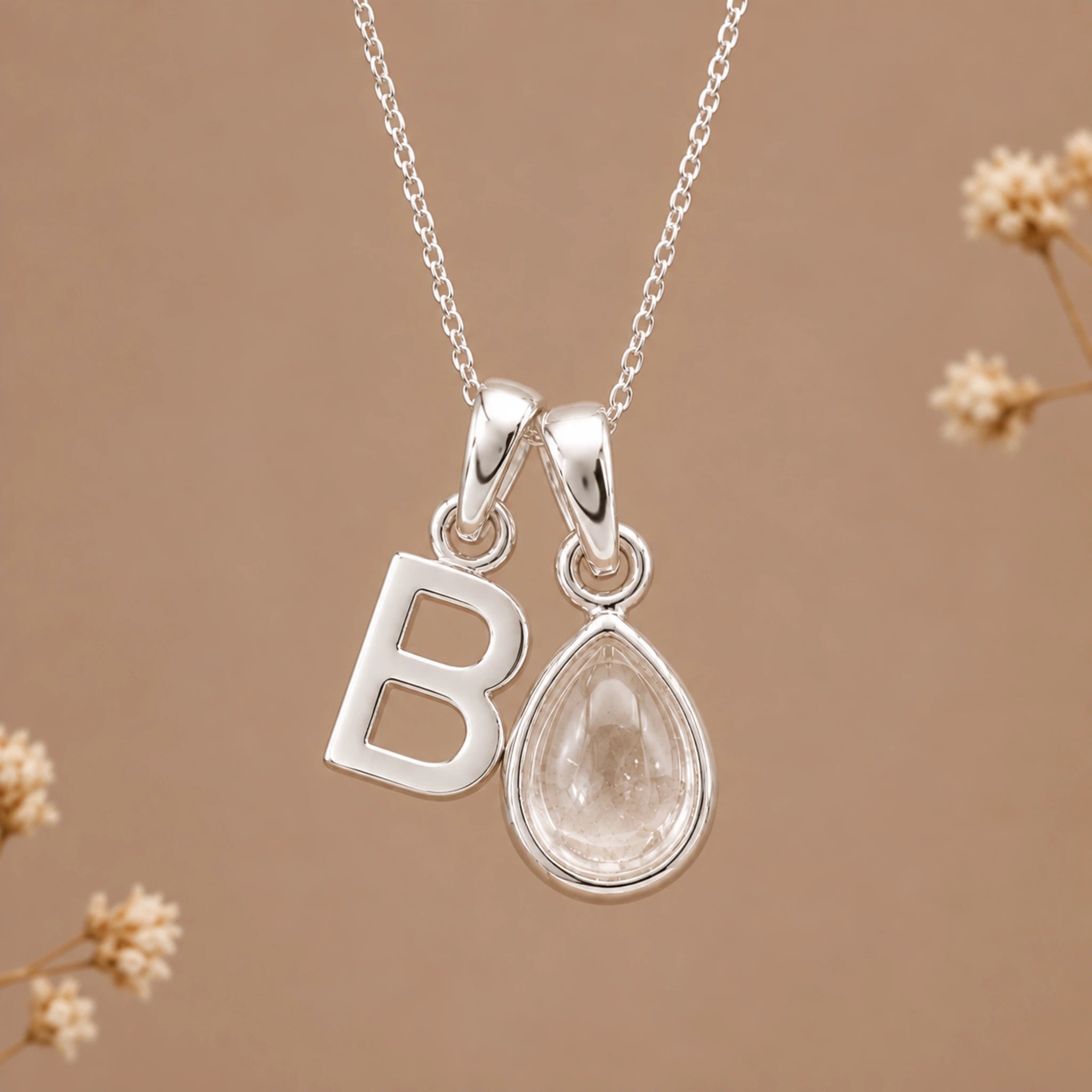 A silver initial necklace featuring a polished letter 'B' pendant and a teardrop-shaped clear gemstone pendant, hanging from a delicate silver chain against a warm beige background with subtle dried floral accents.