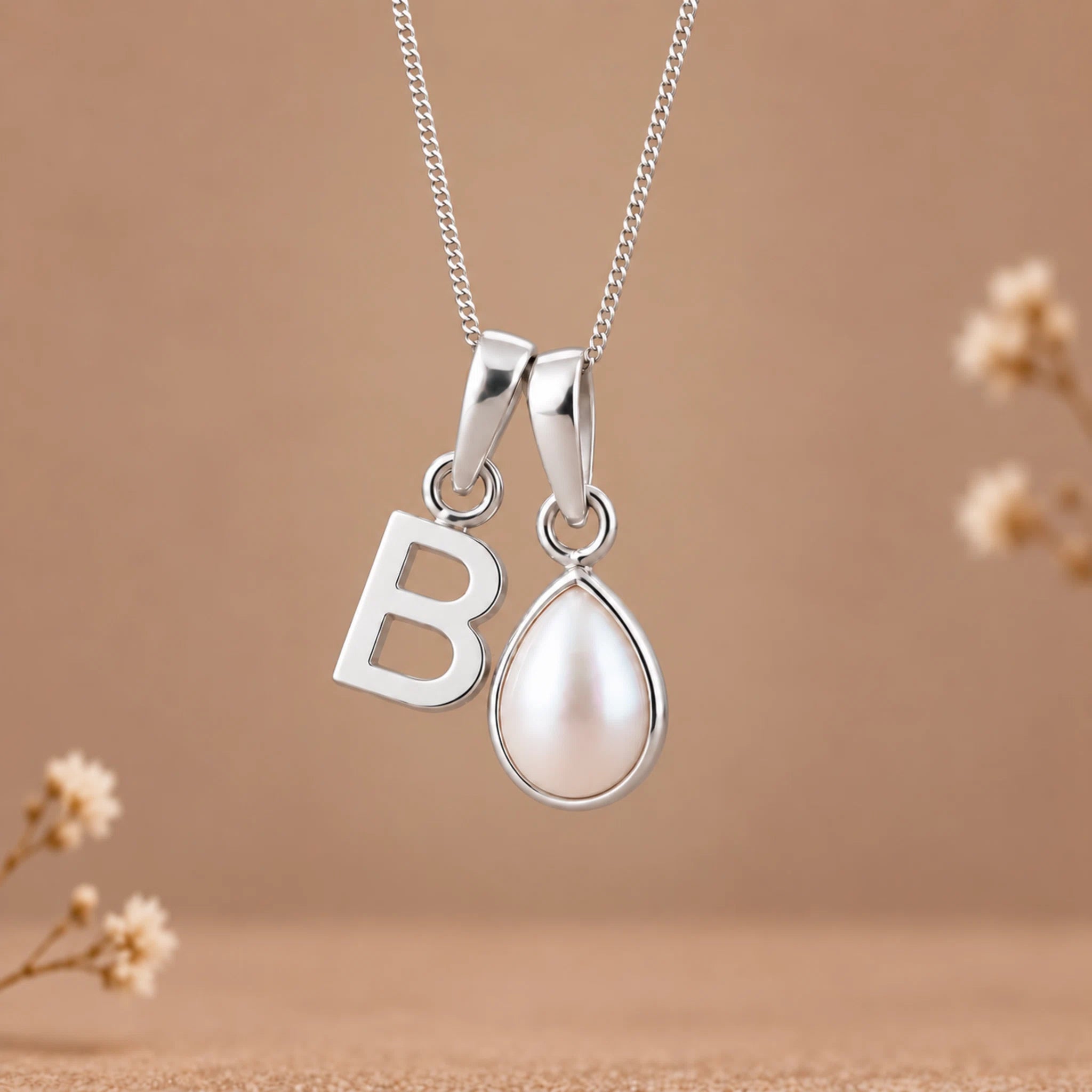 A silver initial pendant necklace featuring a cutout letter 'B' and a teardrop-shaped pearl pendant, displayed against a warm beige background with subtle dried floral accents.