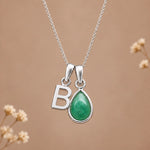 A silver initial pendant necklace featuring a teardrop-shaped green stone and a polished silver letter 'B', displayed against a warm beige background with subtle dried floral accents.
