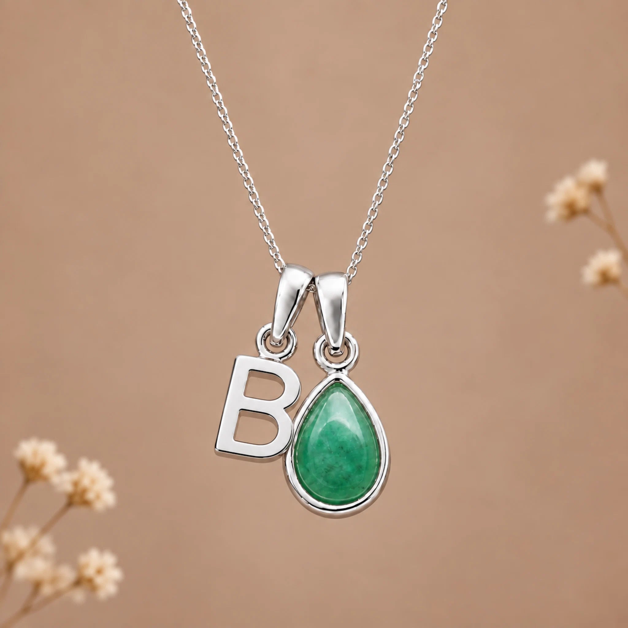 A silver initial pendant necklace featuring a teardrop-shaped green stone and a polished silver letter 'B', displayed against a warm beige background with subtle dried floral accents.